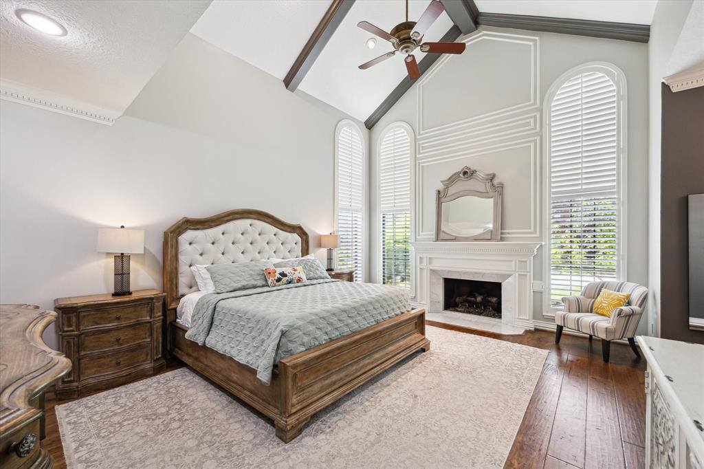 This elegant bedroom features a high ceiling with a ceiling fan, large arched windows with plantation shutters, and a cozy fireplace. The room is furnished with a tufted headboard bed, wood furniture, and accent lighting, creating a warm and inviting atmosphere.